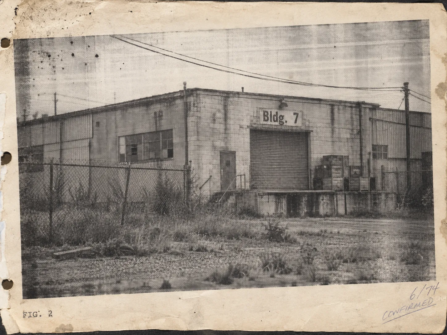 Photocopied black-and-white photograph of a concrete warehouse marked Bldg. 7 behind a chain-link fence